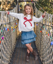 Load image into Gallery viewer, A young girl stands on a suspension bridge wearing a white knit sweater with a red bow graphic, a blue denim skirt, and brown boots. The bridge has rope railings decorated with colorful Christmas lights.
